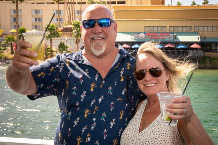 Smiling couple toasting with drinks by a waterfront, wearing sunglasses.