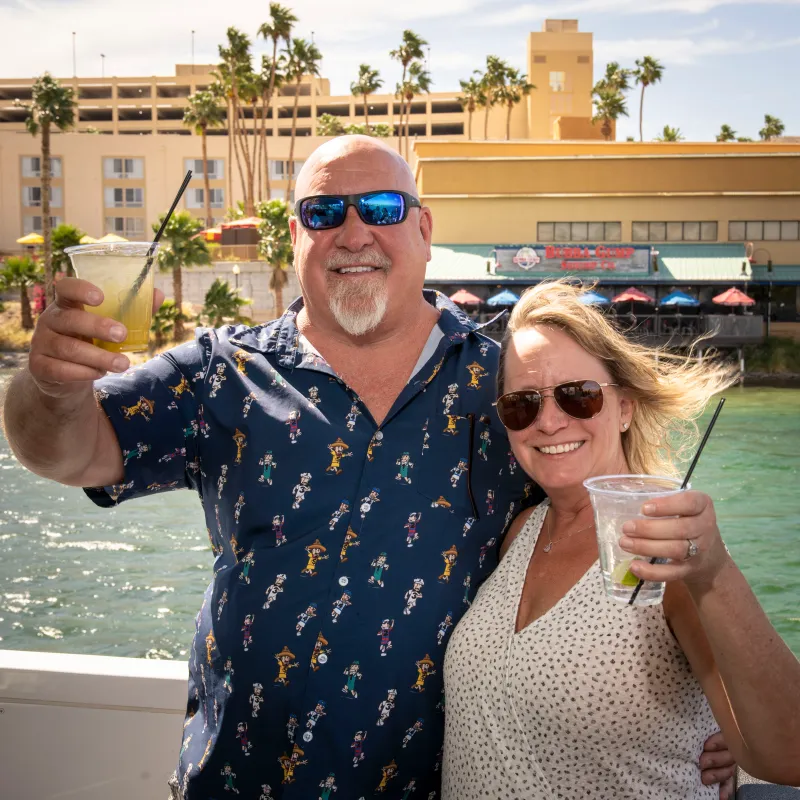 Smiling couple toasting with drinks by a waterfront, wearing sunglasses.