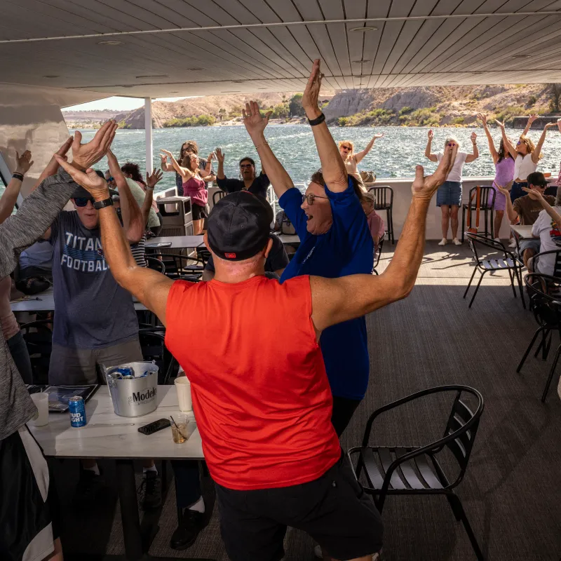 Group of people cheering under a shaded deck near water.