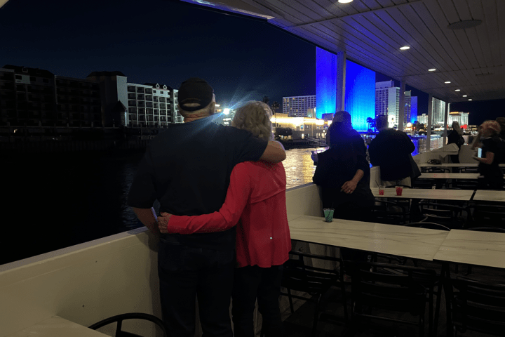 Couple embraces on a boat at night, overlooking colorful city skyline, with tables and other people nearby.