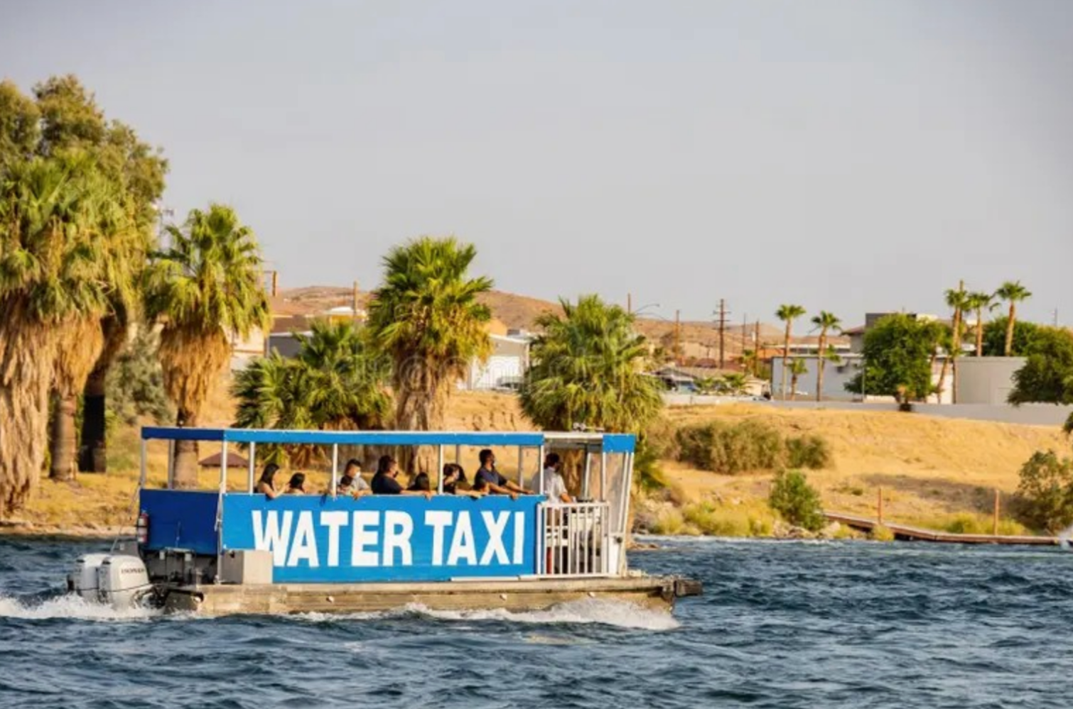 A blue water taxi with passengers on a river near palm trees and desert landscape.