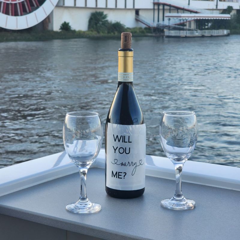 Wine bottle with 'Will you marry me?' label, flanked by two glasses, on a boat deck.