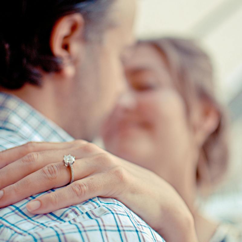 Close-up of hand with engagement ring on shoulder, couple blurred in background.