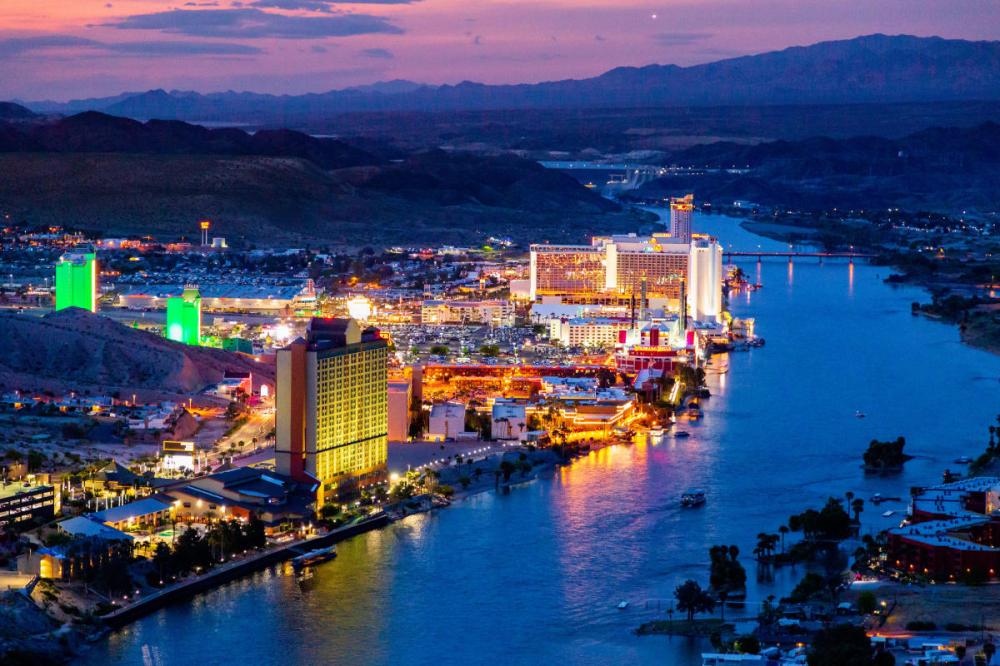 Colorful cityscape at dusk with bright lights along a river under a colorful sky.