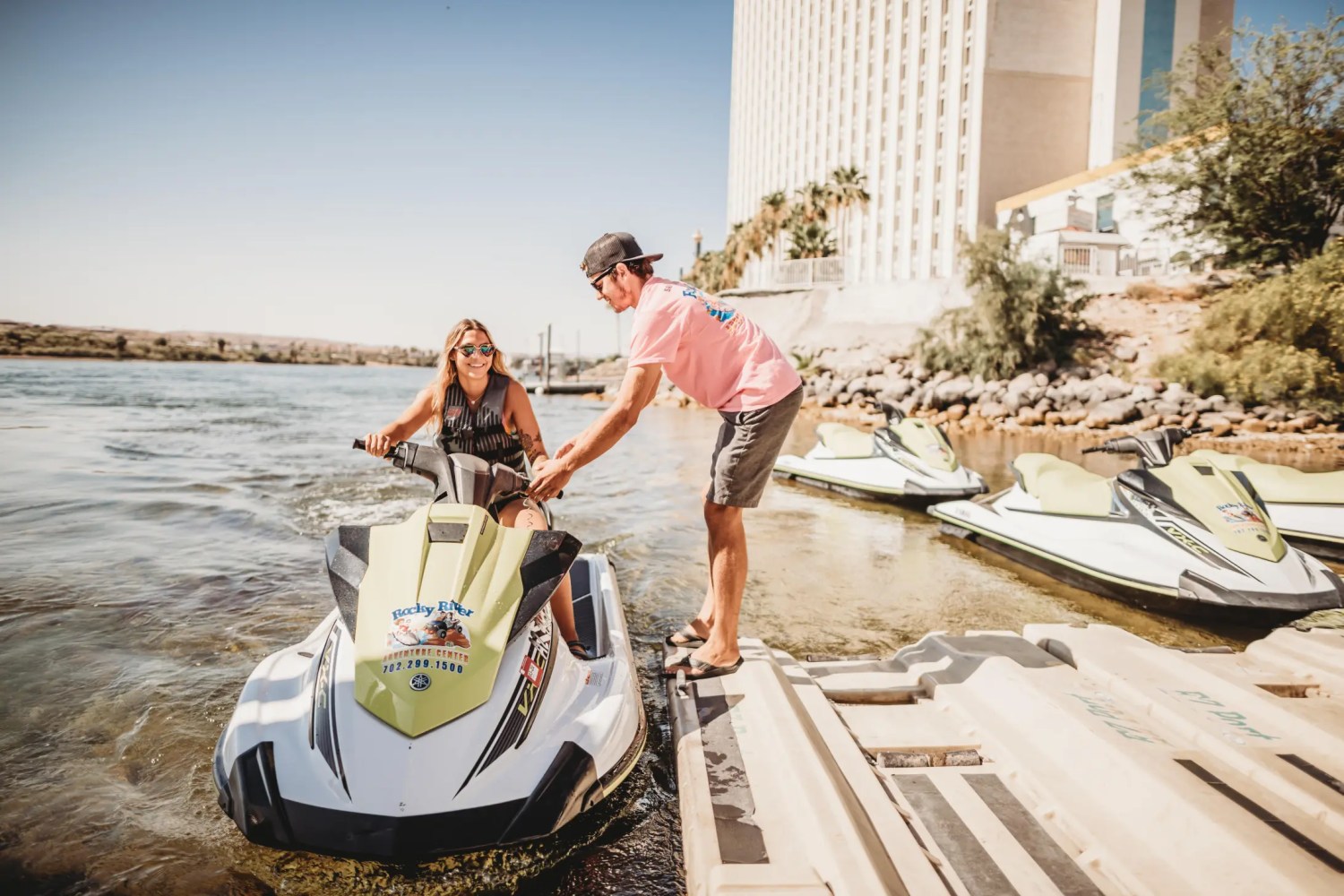 Man helps woman prepare jet ski at riverside dock near large building and clear sky.