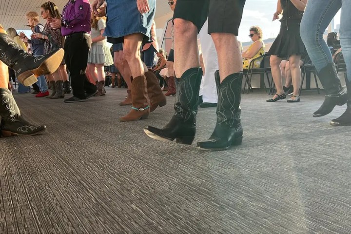 People in cowboy boots line dancing indoors on a carpeted floor.