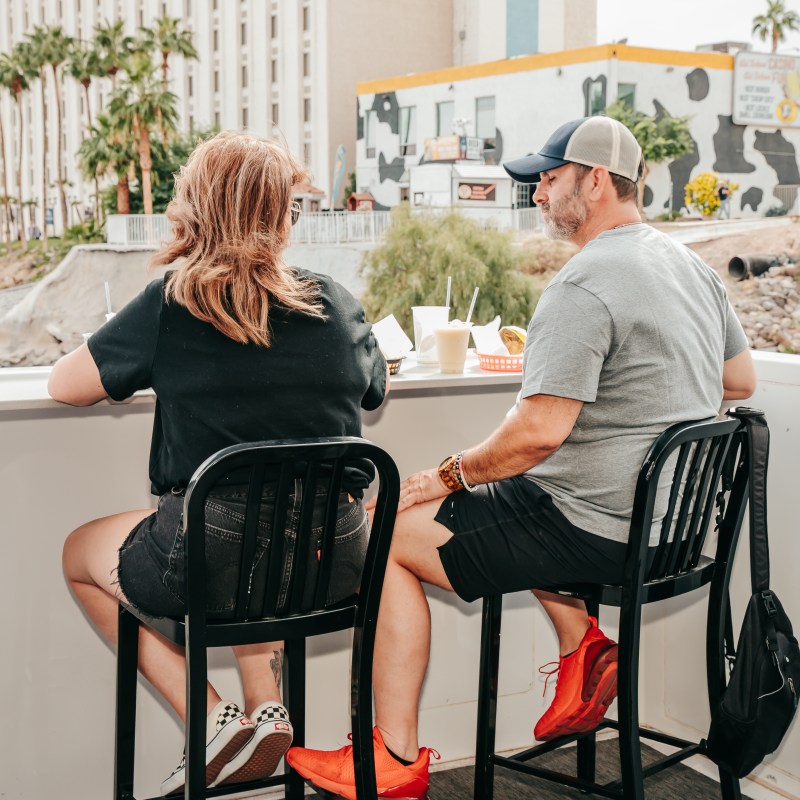 Two people sitting outside at a bar, view of a city building and trees in background.
