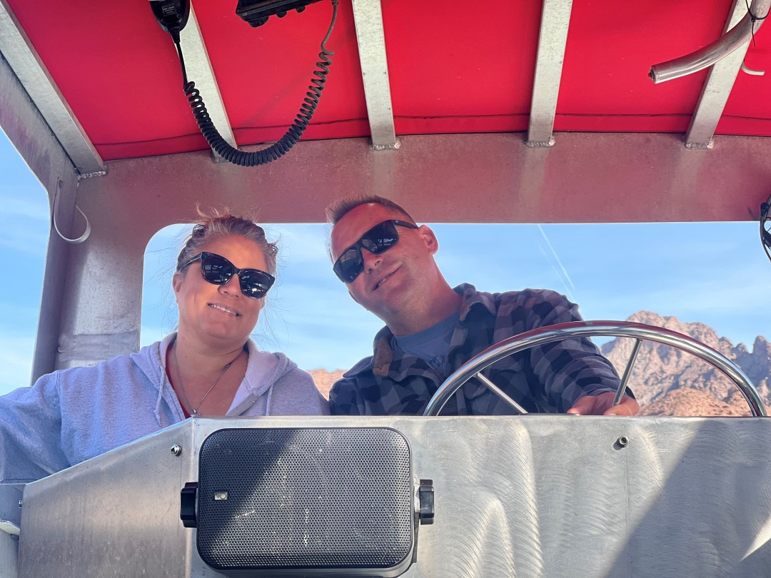 Two people wearing sunglasses steering a boat with a red canopy.